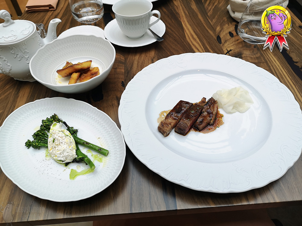 On the table is a large plate with sliced beef and kohlrabi flakes. Next to it is a platter with grilled wild broccoli and burrata and a white bowl with pieces of oven potatoes partially dipped in buttermilk.