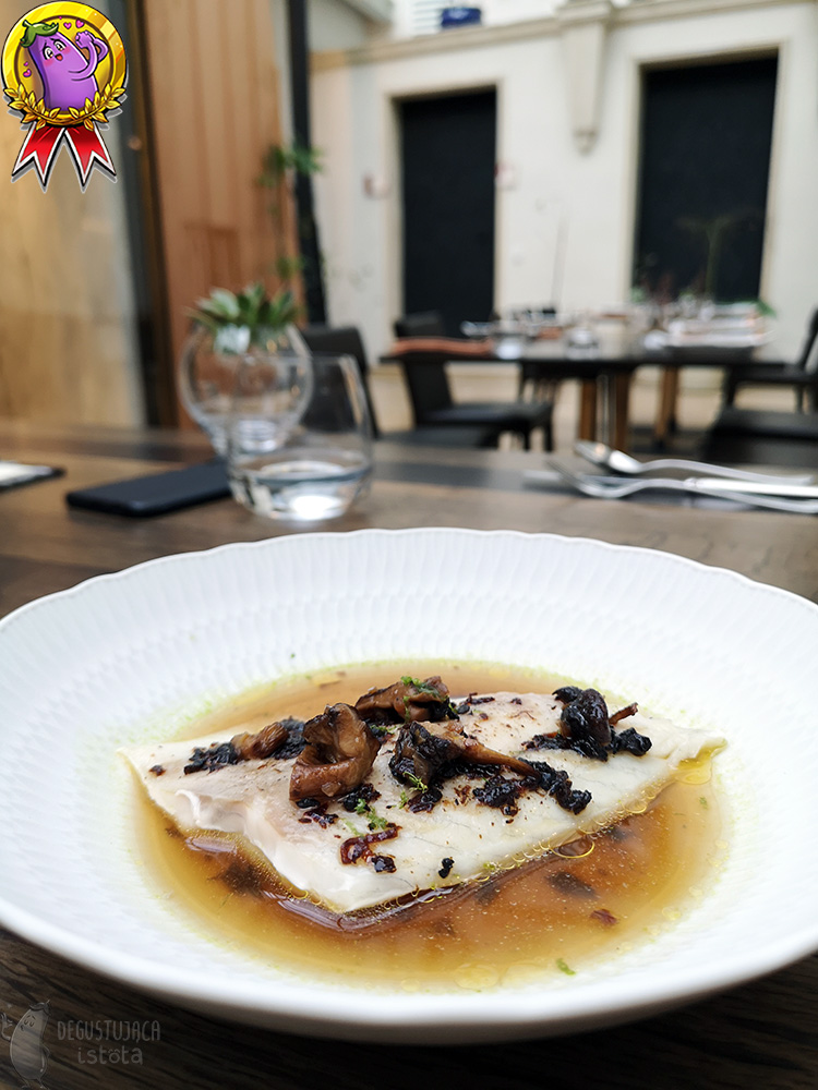 A piece of perch without skin is in a broth in a deep, white plate. Topped with pieces of shiitake mushrooms.