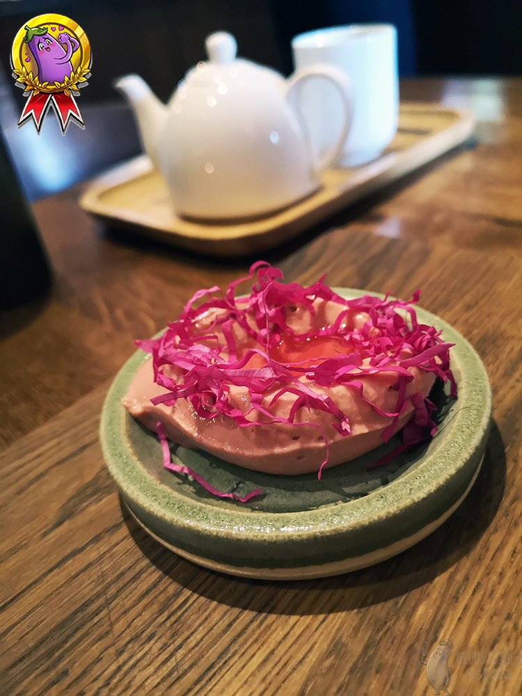 Różowe lody udekorowane paskami z płatków róż i oblane syropem różanym. Całość ułożona na małym, szarym talerzyku.