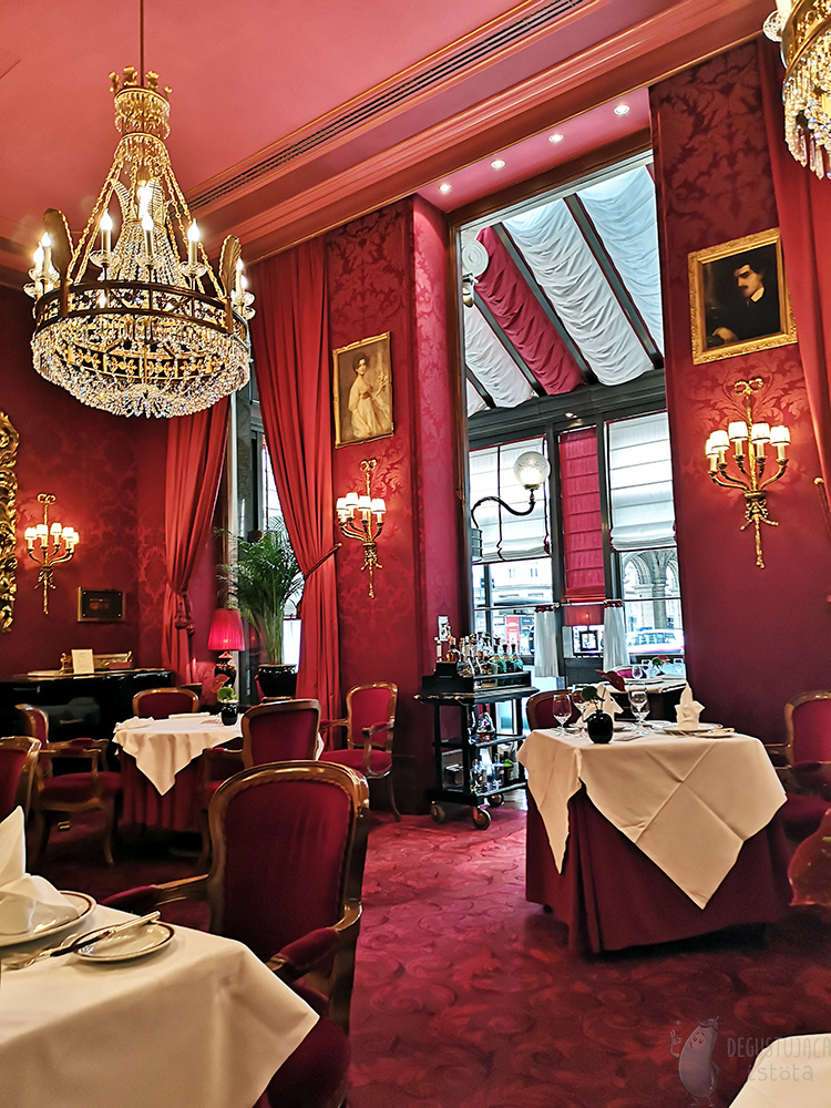 A view of the center of the restaurant room. A crystal, gold-plated chandelier hangs from the ceiling. The carpeting on the floor is also red.