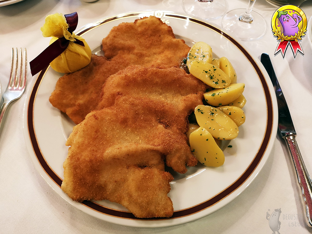 On a large flat plate lie two schnitzels, one on top of the other. To the right of the schnitzels are halves of potatoes, poured with fat and sprinkled with parsley. On the left side there is a lemon half wrapped in a yellow net, tied with a maroon ribbon.