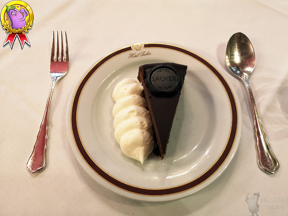 A piece of chocolate cake with a portion of whipped cream on a small white plate. Next to it there is silver cutlery. Top view of the cake.