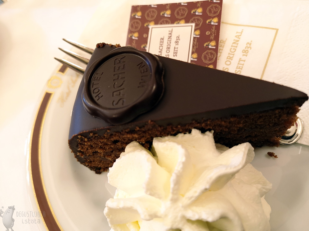 Kawałek czekoladowego torcika z porcją bitej śmietany na białym, małym talerzyku.