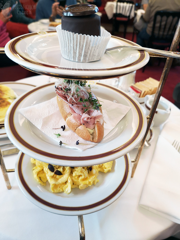 Set of three plates on stand. On the top one is a Sacher Punch Dessert in paper. On the middle one, a sliced roll filled with ham and sprinkled with horseradish and cuckooflower. On the last, a portion of scrambled eggs with pumpkin seeds placed on top.