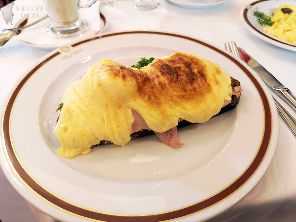 A slice of bread doused with top-baked hollandaise sauce. Ham sticks out from under the sauce. The bread lies on a white flat plate with the logo of Hotel Sacher.