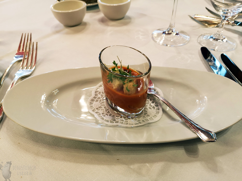 On an oval white plate, on a small, white paper napkin lies a small glass filled with red gazpacho. Decorated on top with tiny croutons and cuckooflower.