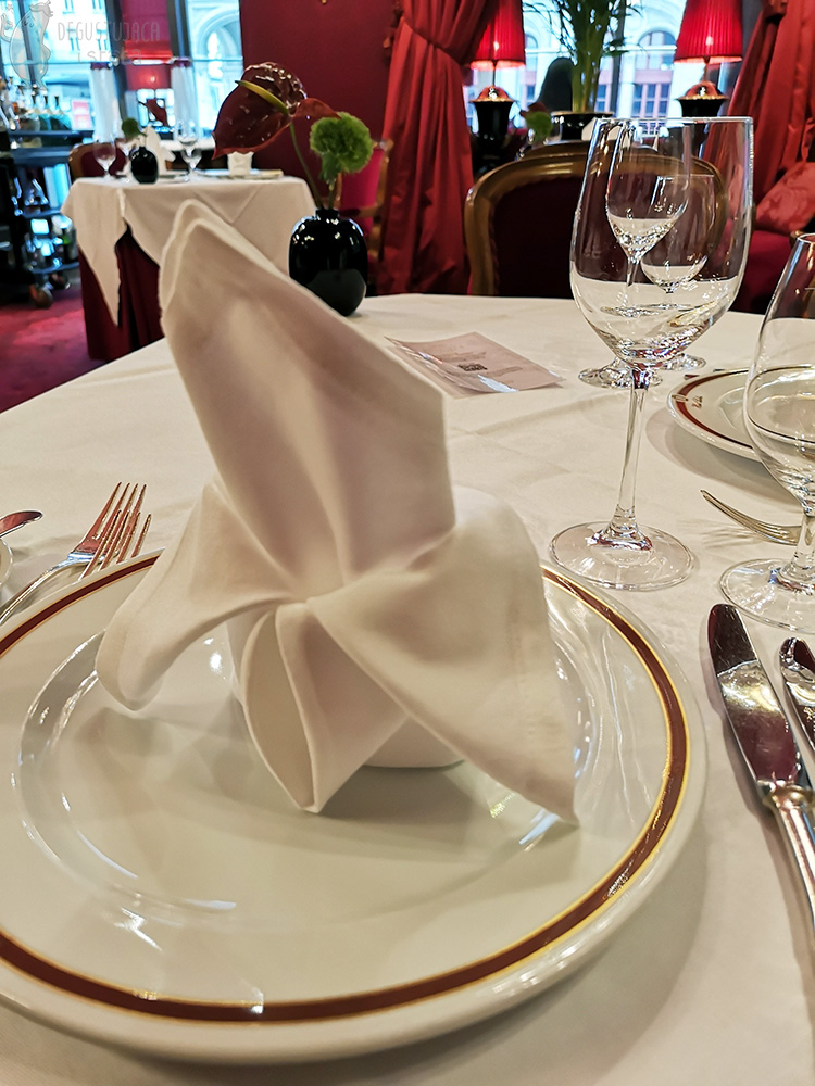 Pięknie złożona biała serwetka położona na talerzu z logiem Hotelu Sacher. Obok położone są srebrne sztućce.