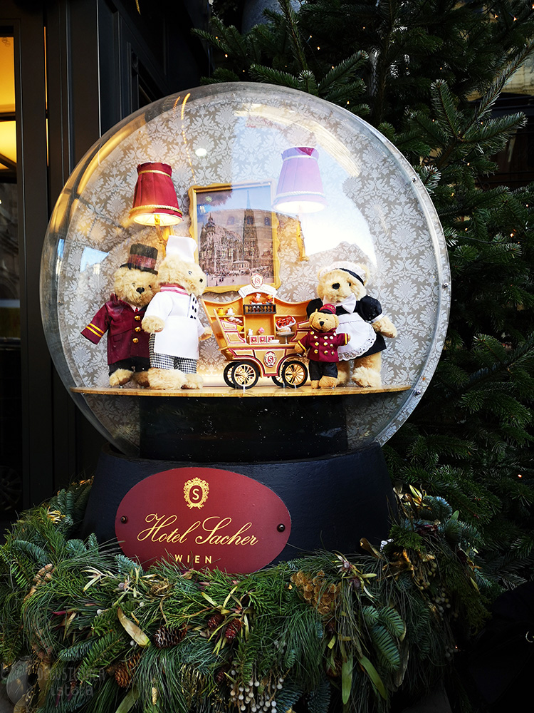 Large transparent ball with three big teddy bears and one little bear. One big bear sells sweets from his stall on wheels. The ball stands on a black stand with the Sacher Hotel logo.