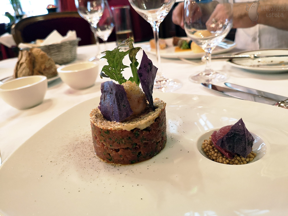 On the white plate to the left is tartare, arranged in a disc, smeared on top with mayonnaise, garnished with green and purple leaves. On the right, a portion of mustard and grape ice cream is arranged in the spherical hollow of the plate.