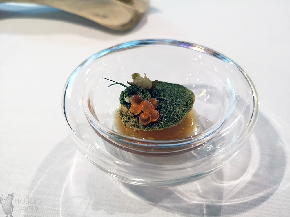 In a transparent glass bowl, there is a disk sprinkled with dill powder, orange mousse underneath and orange roe on the disk.