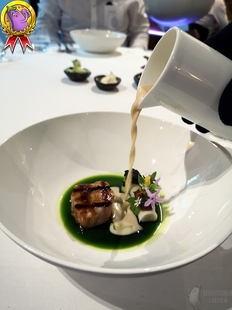 In a deep white plate, whose bottom is covered with green oil, a piece of grilled fish is arranged. Next to the catfish there are white pipes decorated with flowers and caviar. From a white jug is poured white, smooth sauce.