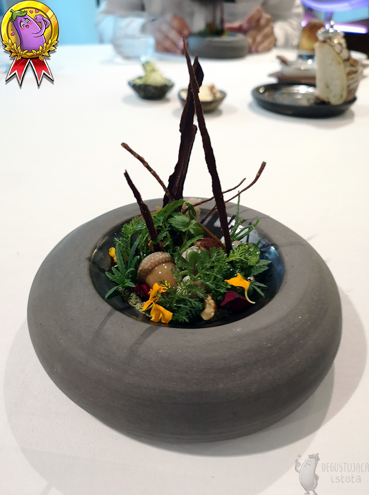 In a thick, dark grey dish arranged There are mushrooms and acorns from liver mousse, surrounded by green twigs, yellow and burgundy flower petals, and vertically arranged pieces of chocolate.