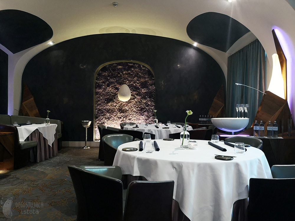 Room with large round tables covered with white tablecloths. On the central, black wall there is a large amethyst block, illuminated from underneath with a hanging white egg.