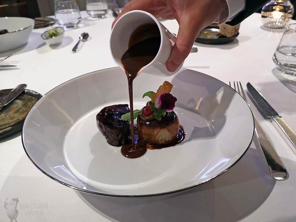 On a flat white plate On the left is a dark piece of lamb, and on the right is an onion decorated with leaves and cut out roses. In the middle a dark sauce is poured.