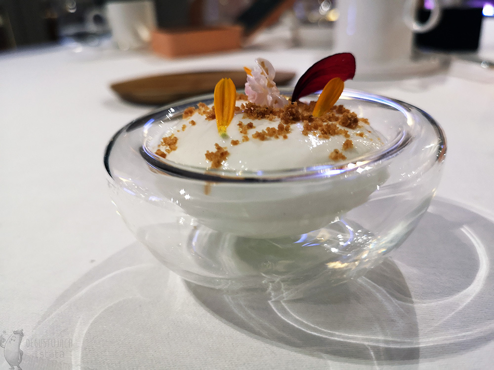 In a glass bowl there is The foam is decorated with crumble and petals of orange flowers from the top and a small pink rose.
