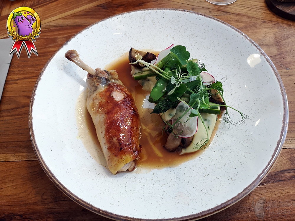 On a bright, flat plate lies a stuffed guinea fowl's leg. Next to it is a portion of puree with green asparagus, mushrooms and sugar peas shoots on top. The whole is poured with baking sauce.