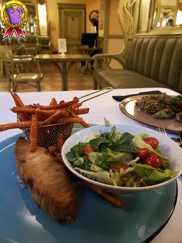 On a flat, blue plate lies a coated piece of halibut and in a melted basket of sweet potato fries. On the plate there is also a bowl with a salad with cocktail tomatoes.