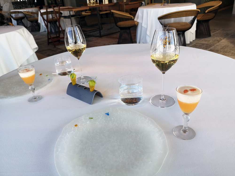 Zdjęcie stołu z białym obrusem, na którym leżą dwa duże talerze, kieliszki z szampanem oraz kieliszki z pomarańczowym pisco sour. Na środku stołu w podstawce umieszczone są dwa rożki z zielonym nadzieniem.