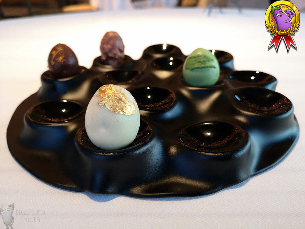 Czarny talerz z miejscami na jajka. Umieszczone są tam 4 małe jajka: dwa brązowe, zielone i białe maźnięte złotem.