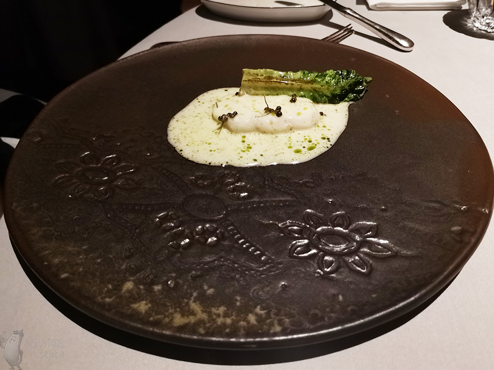 W rogu ciemnobrązowego talerza położony jest kawałek sandacza, oblany białym sosem. Obok grillowany liść buraka pod którym ukryte są szparagi.