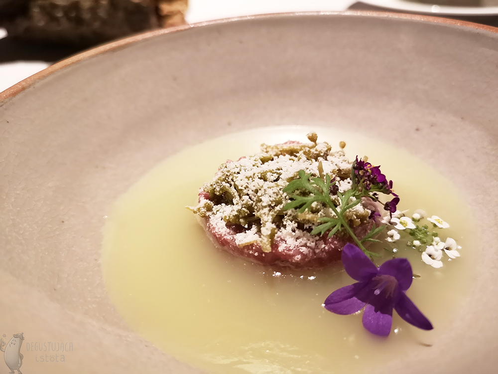 A bowl with yellow liquid inside a tartare disk is laid, sprinkled with white horseradish powder and decorated with white, small flowers and one purple bell.