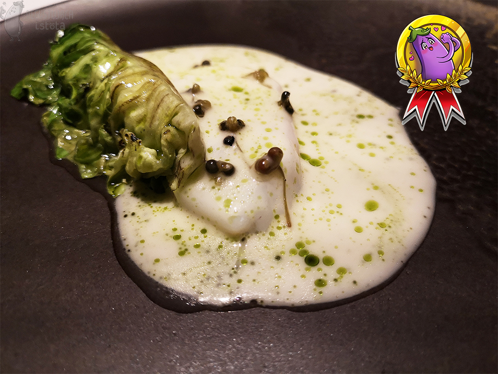 Kawałek białej ryby polanej białym sosem z zielonymi kroplami oliwy, podany na karnym talerzu. Obok zwinięty, przygrillowany liść buraka cukrowego.