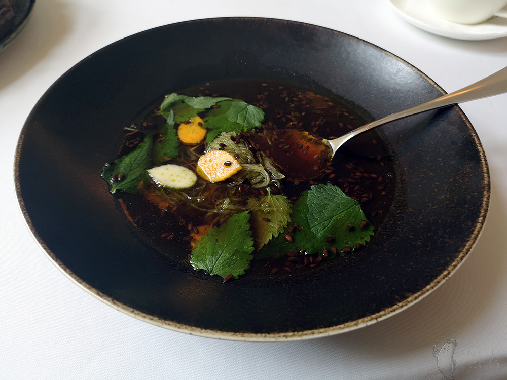 W ciemnym talerzy rozmieszana już zupa w której widać pomarańczowe i zielonkawe listki wycięte z warzyw oraz zielony makaron. Na powierzchni zupy pływa siemię lniane.