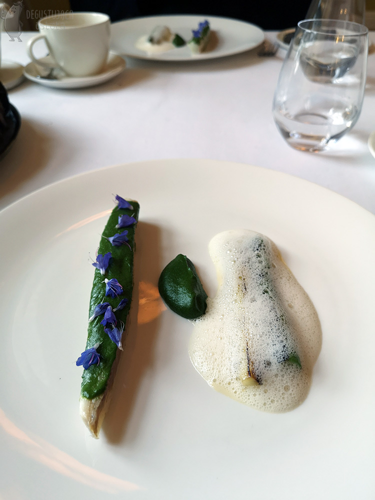 Na płaskim białym talerzu po lewej leży pasek okonia posmarowany z wierzchu puree szpinakowy i ozdobionym niebieskimi kwiatkami. Na środku umieszczona jest porcja tego samego puree, a po prawej biały szparag i por przykryte pianą z masła. 