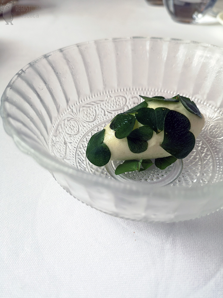 Na szklanym talerzyku położony jest biały sorbet obłożony zielonymi listeczkami.