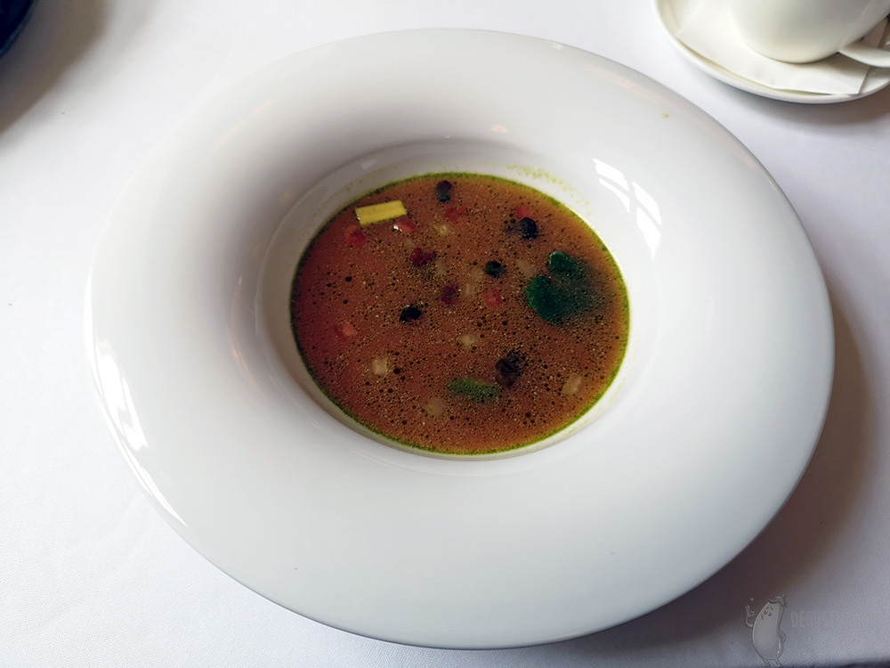 W białym talerzu jest czerwono żółta zupa z zieloną otoczką i pływającymi w niej warzywami.