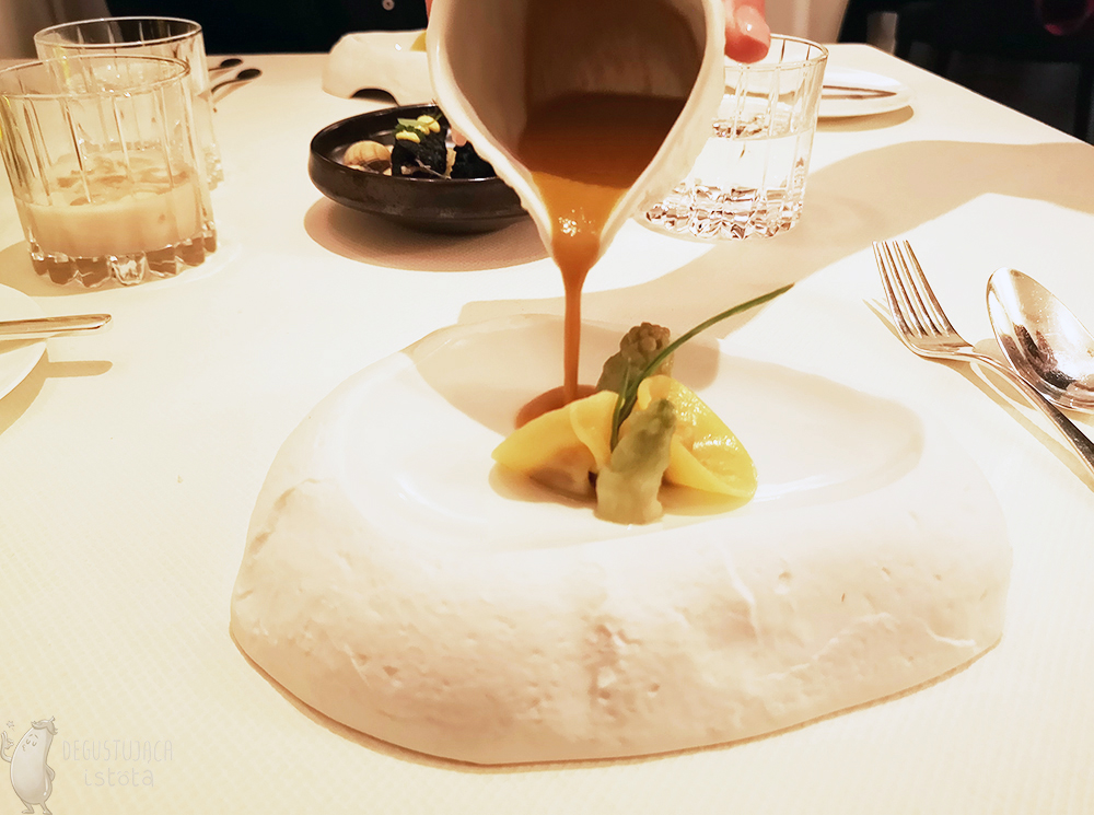 Biały kamień wydrążony w kształt talerza, a na nim: dwa kawałki zielonych czubków Cicoria Asparago, obok dwa ravioli i sos w trakcie nalewania. Na środku stołu widać opanierowane sardynki na talerzu, położone na uszlach