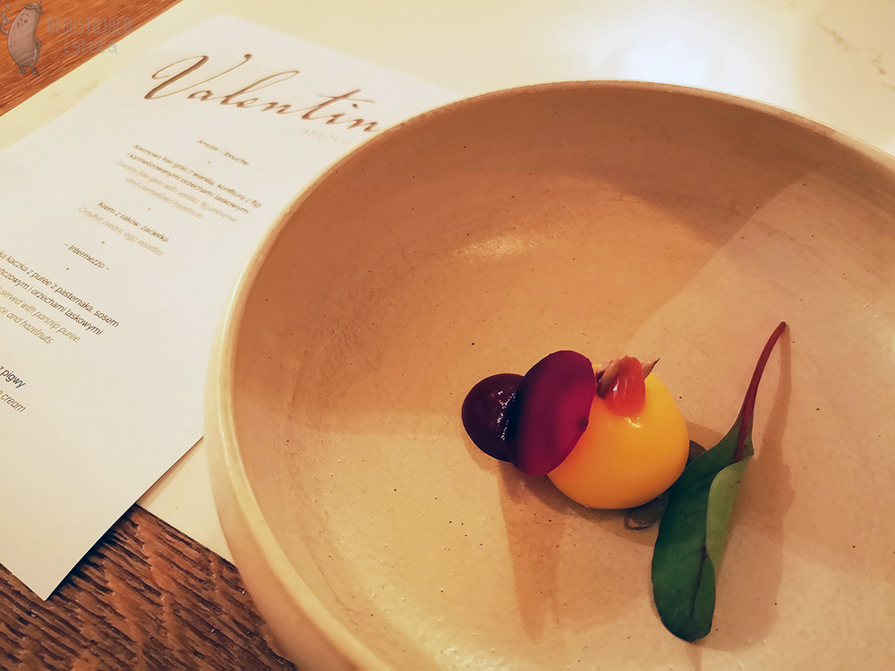  A bowl with a yellow ball made of goat cheese, next to which lies a dot of beetroot mousse and a thin slice of beetroot.  The whole dish is decorated with pumpkin seeds and beet leaf.
