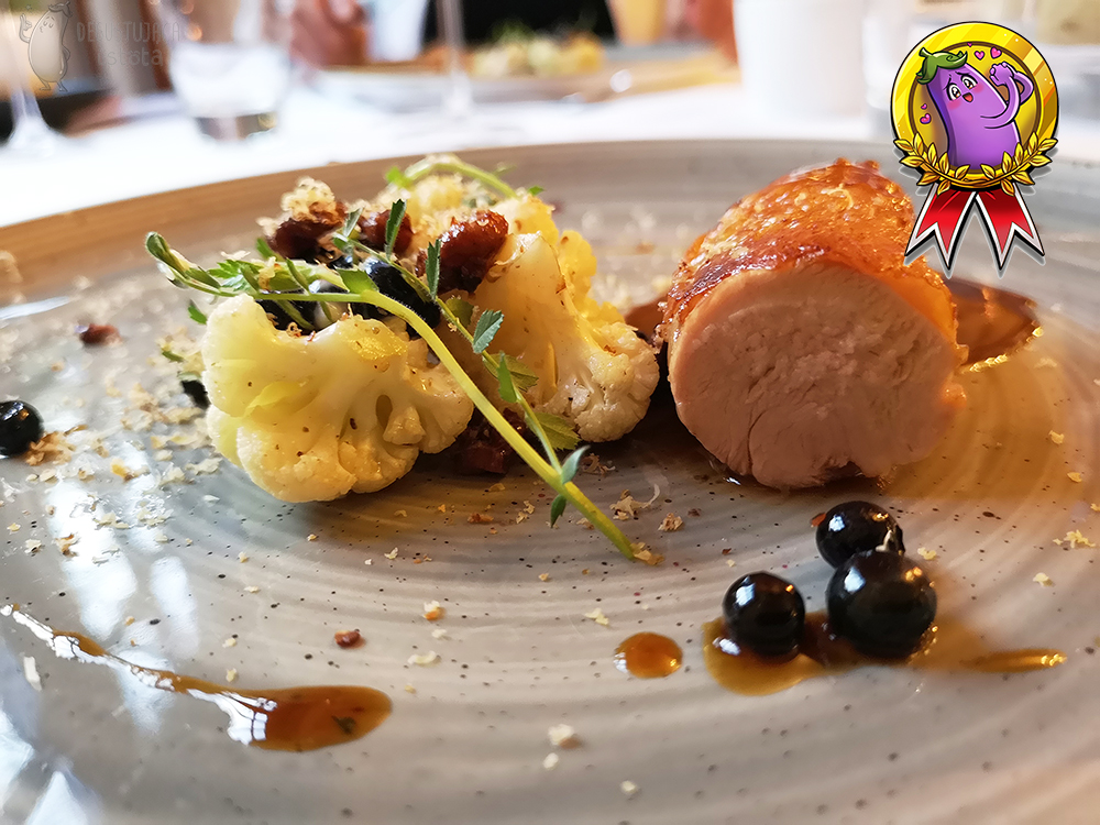 A round piece of guinea fowl meat with toasted skin, next to pieces of cauliflower and a few berries. Served on a gray plate and drizzled with roast sauce.