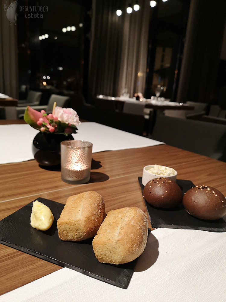  Dwa czarne kafle, położone na stole, na których są umieszczone po dwie sztuki, mini chlebków i ciemnych okrągłych bułeczek. Przy chlebkach nałożone jest masło a przy bułeczkach w białej miseczce, smalec.