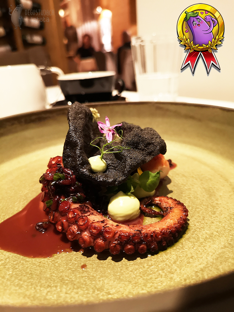 A piece of octopus tentacle with a black chip and slightly greenish mayonnaise applied. Decorated with a small pink flower.