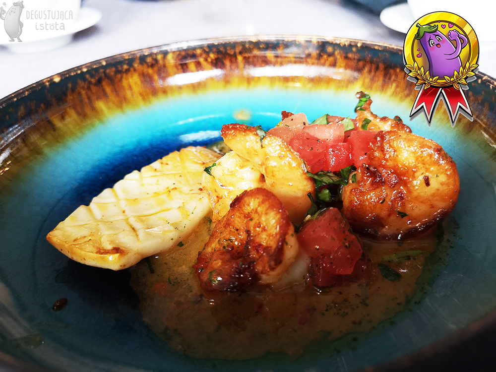 Kawałek kalmara, przysmażonego i naciętego w kratkę, obok przysmażone krewetki z kawałkami pomidorów na błękitnym talerzu, brązowym po bokach.