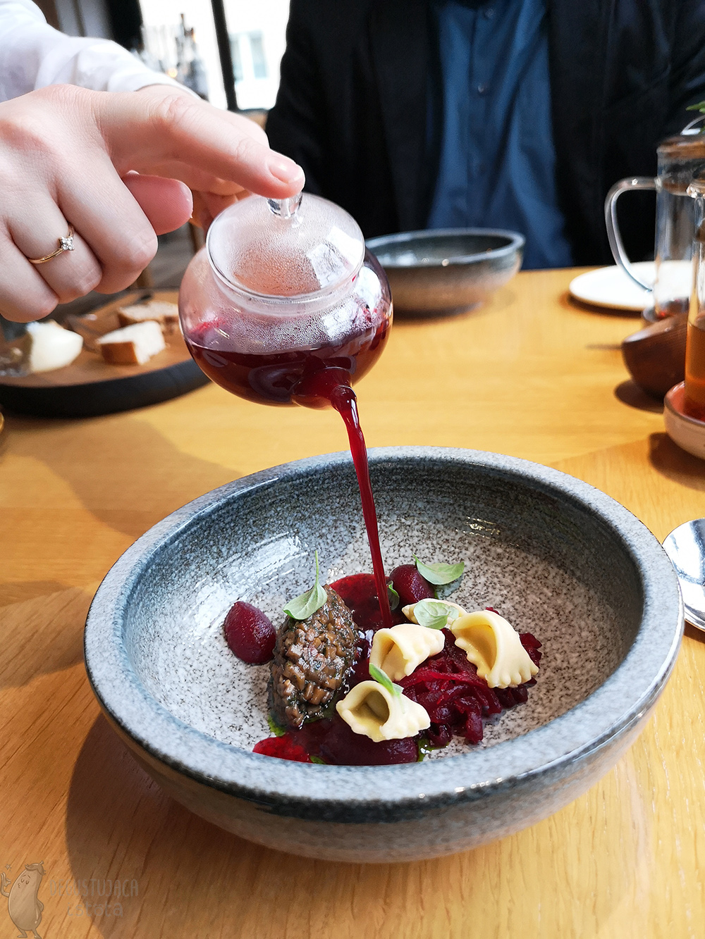 Borscht during pouring from a glass teapot into a stone bowl with small ravioli, beetroot and mushrooms.