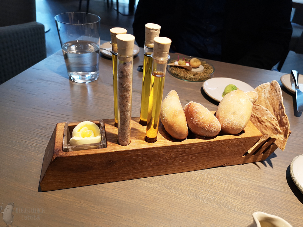  On a wooden, rectangular base are placed 4 glass vials, rose-shaped butter putted into a glass container recessed in the base. Small rolls are arranged behind the vials, and on the end there is crispbread.
