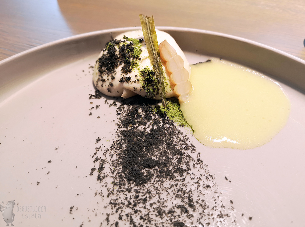  White cream dome on a gray plate, sprinkled with black and green powder. On it lies a piece of glassy, ​​beige crosne next to which a yellow, buttered sauce is poured.
