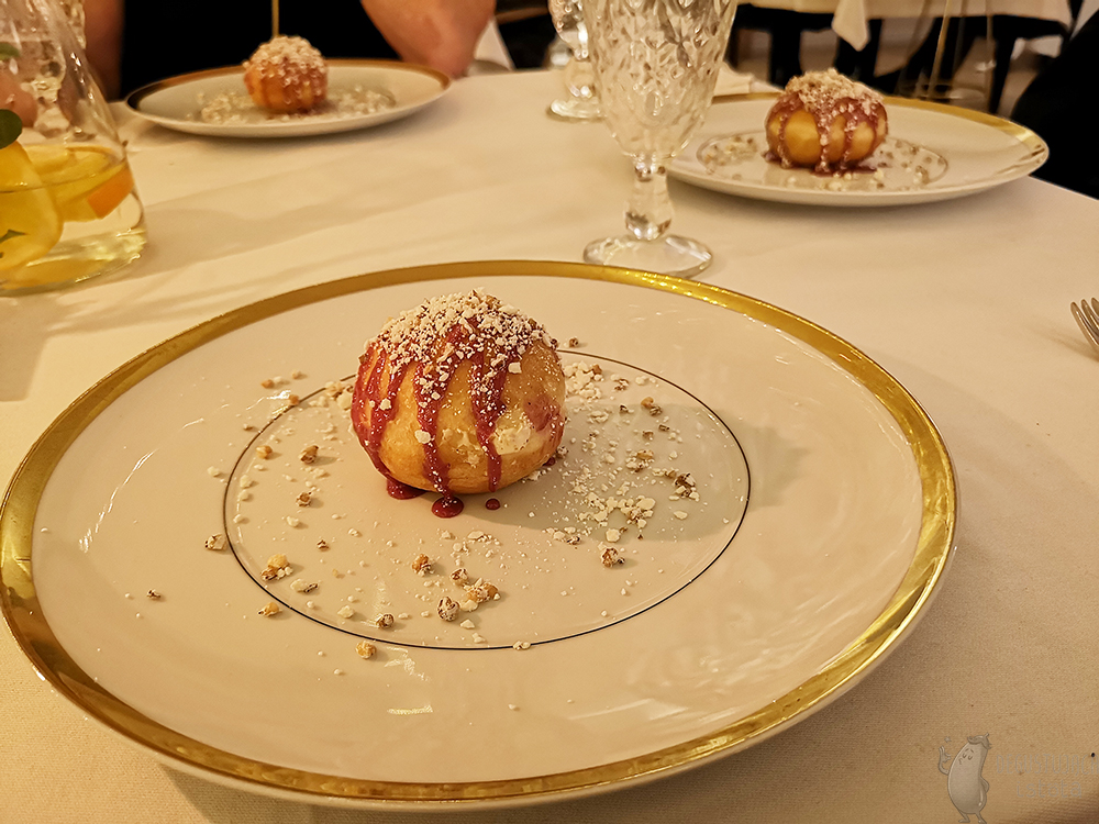 Stół z płaskimi, dużymi talerzami ze złoconym brzegiem na których podane są pączki z różowym lukrem, białym pudrem i popcornem.