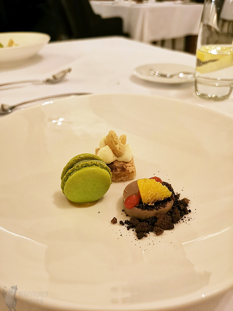 On the plate sweets were arranged. Almond cake, green macaroon and round chocolate mousse with a piece of tangerine.