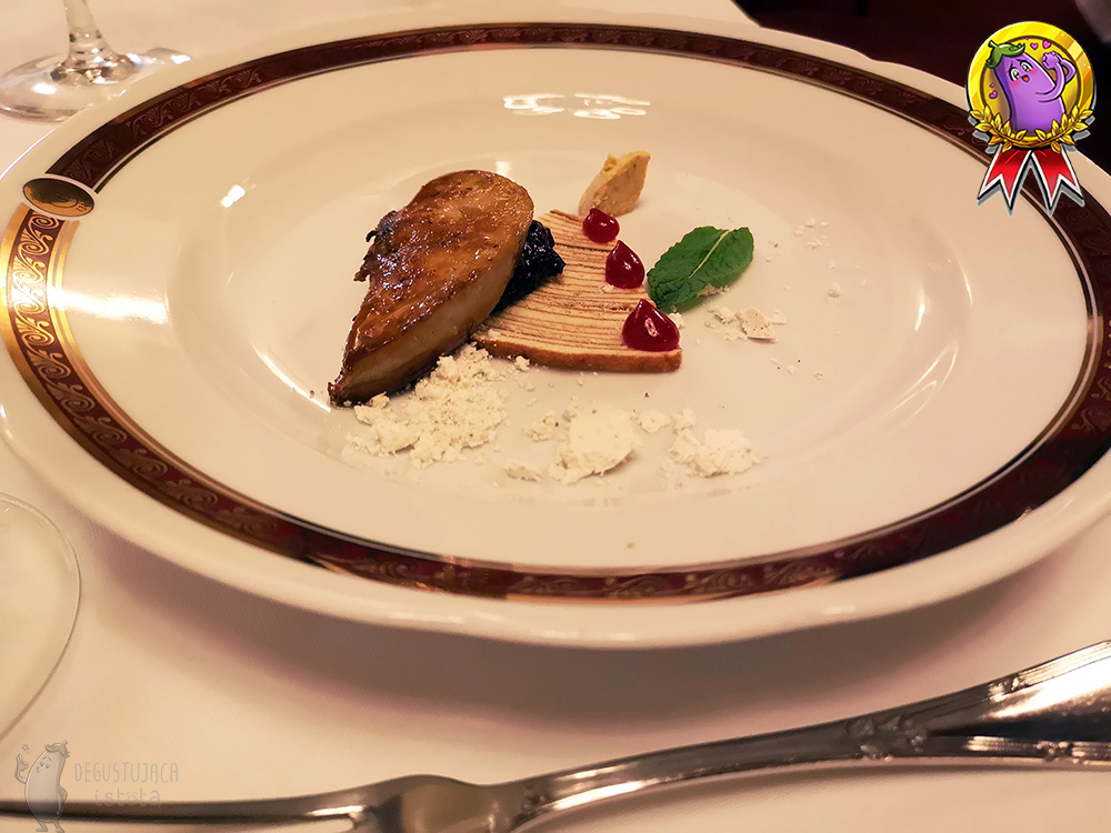 Kawałek przypieczonej wątróbki, leżący na płaskim kawałku sękacza. Obok foie gras, na sękaczu znajdują się czerwone kropki sosu.