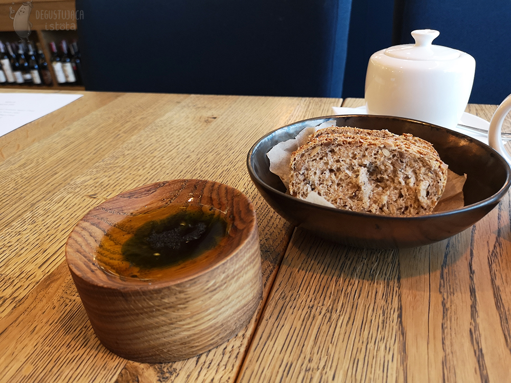 Miska z kromkami ciemnego pieczywa i oliwa podana na wyżłobionym kawałku drewna.